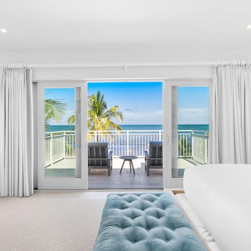 A bright, modern bedroom with a view of the ocean through glass doors, leading to a balcony with palm trees and patio chairs.