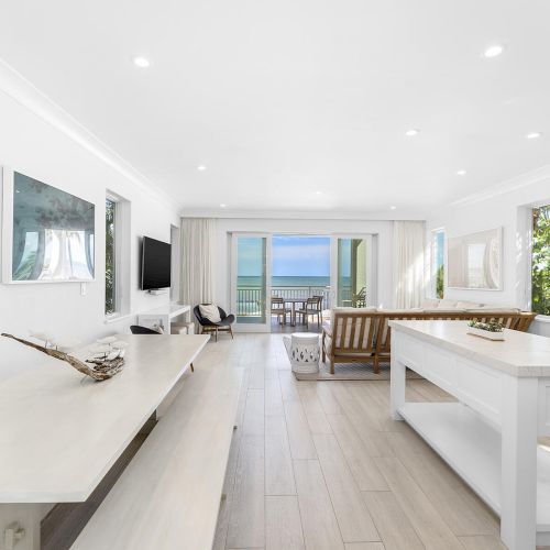 A bright, modern open-concept living room and kitchen with white decor, wood accents, and a view of the ocean through glass doors.