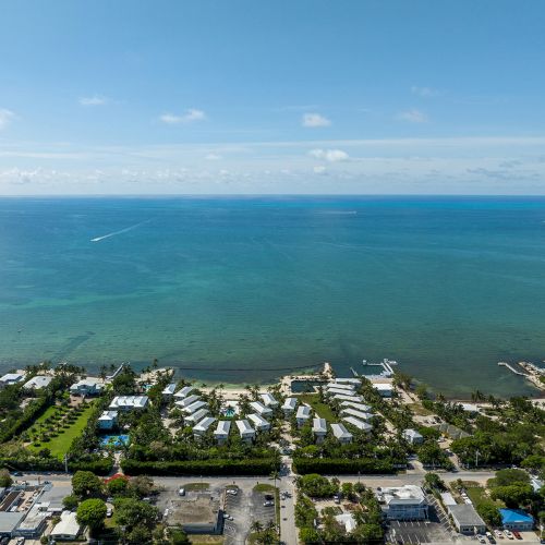 A coastal aerial view with buildings, greenery, and a vast ocean under a clear blue sky, featuring a distant boat creating a white wake.