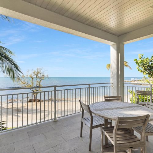 A coastal patio with a wooden table and chairs, overlooking a sandy beach and the ocean with palm trees and a clear blue sky.