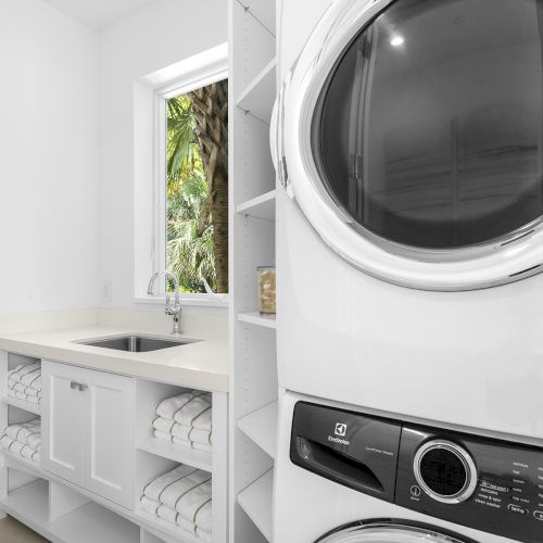 This image shows a laundry room with a stacked washer and dryer, sink, window, and shelves storing folded towels.