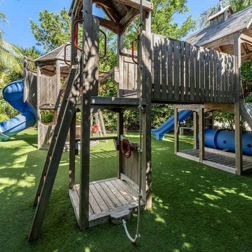 A playground with a wooden play structure, including slides and tunnels, surrounded by trees and greenery, on a sunny day.