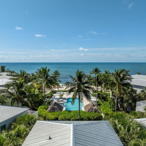 A tropical resort with white-roofed buildings, palm trees, and a pool, facing a clear blue ocean under a sunny sky.