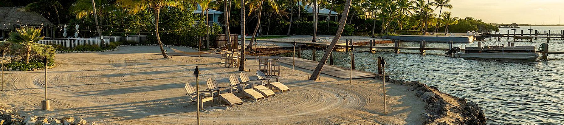 A serene beach setting with palm trees, lounge chairs, and a docked boat at sunset on a rocky shoreline.