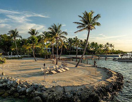 A serene beach setting with palm trees, lounge chairs, and a docked boat at sunset on a rocky shoreline.