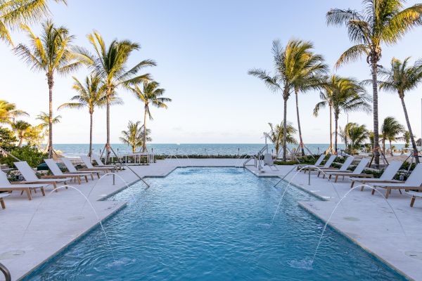 A beachfront swimming pool with lounge chairs and palm trees by the ocean, framed by clear skies and gentle waves.