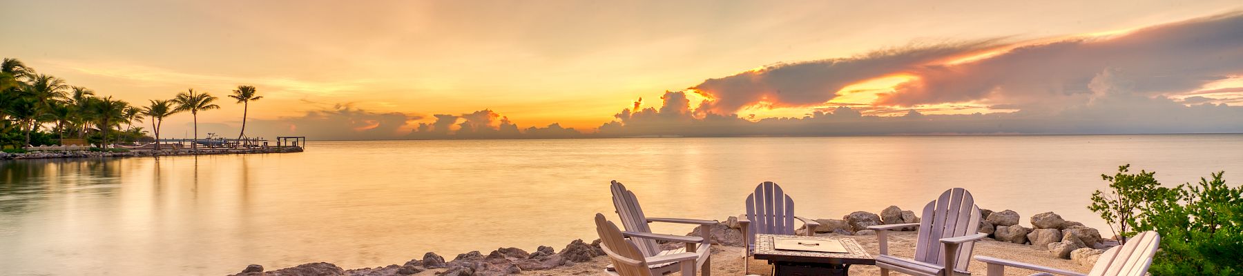 A serene beach setting with Adirondack chairs around a fire pit, overlooking a calm sea at sunset.