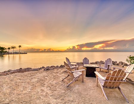 A serene beach setting with Adirondack chairs around a fire pit, overlooking a calm sea at sunset.