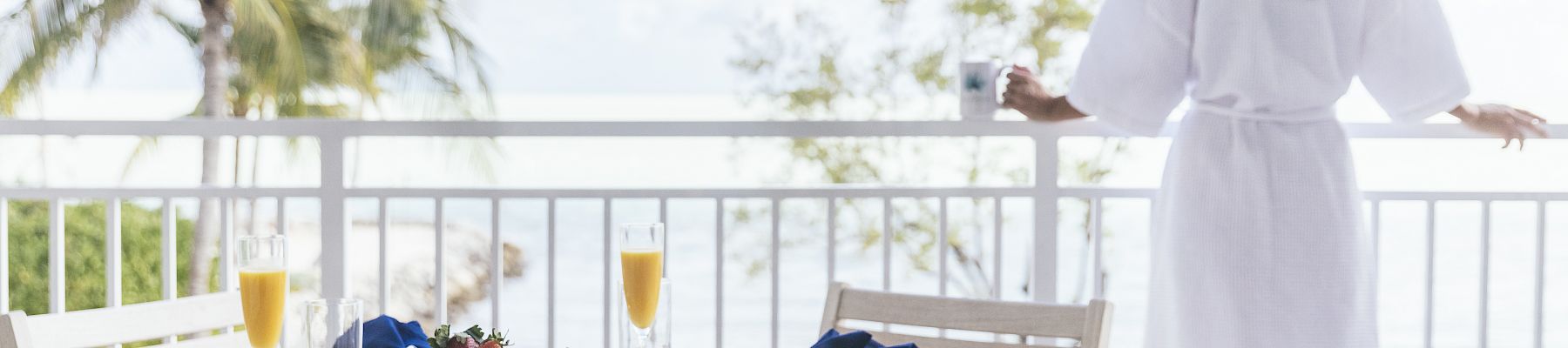 A person in a robe gazes at the ocean from a balcony, with a table set for a meal featuring seafood and mimosas.