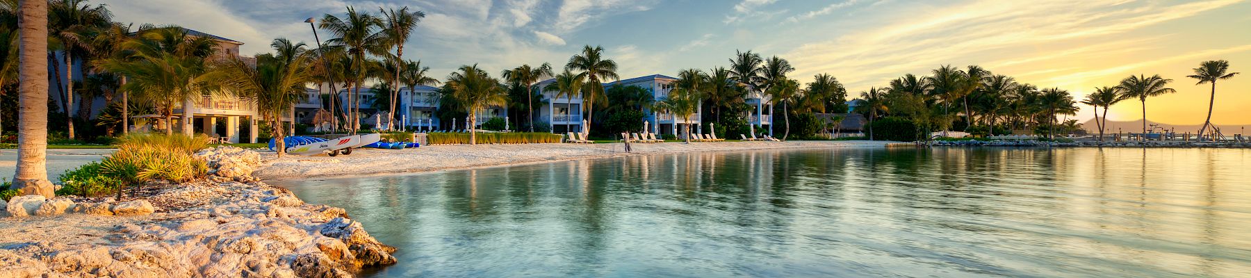 A serene beach scene with palm trees, rocky shoreline, clear water, and a vibrant sunset sky reflecting on the ocean.