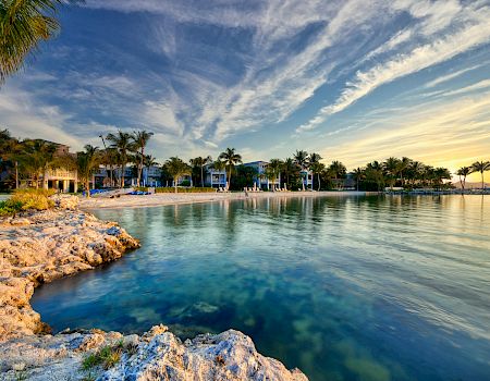 A serene beach scene with palm trees, rocky shoreline, clear water, and a vibrant sunset sky reflecting on the ocean.