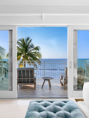 A serene bedroom scene with a view of a palm-lined balcony and ocean, through glass doors, featuring cozy chairs and a footstool.