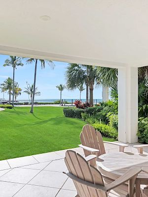 A patio with wooden chairs overlooks a vibrant green lawn and palm trees, leading to a scenic ocean view under a clear blue sky.