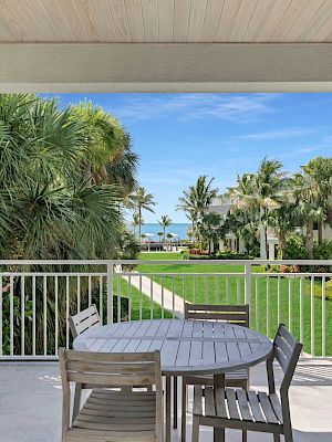 A patio with outdoor furniture offers a view of lush greenery and palm trees leading to the ocean under a clear blue sky.