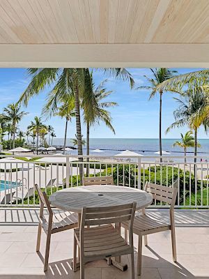 A covered patio with a table and chairs overlooks a pool, palm trees, and a beach with umbrellas under a clear blue sky.