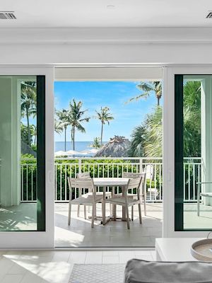 A bright room with white decor opens to a balcony with a table set, overlooking palm trees and a partial ocean view, creating a serene ambiance.