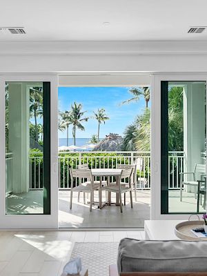 A modern living room opens onto a balcony with a scenic tropical view, featuring palm trees, a table with chairs, and lush greenery.