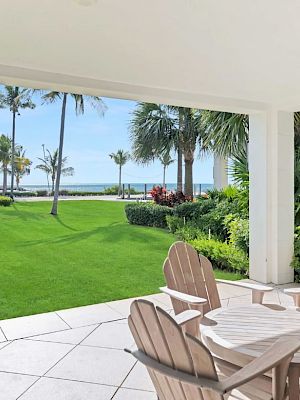 A patio with wooden chairs and a table overlooks a lush garden and the ocean, with palm trees lining the path.