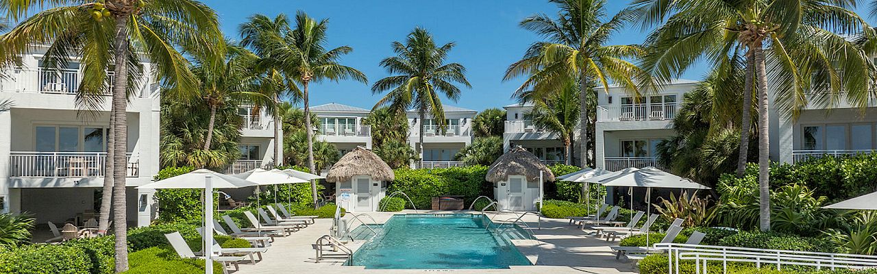 A tropical resort with palm trees, a swimming pool, lounge chairs, and white buildings under a clear blue sky.