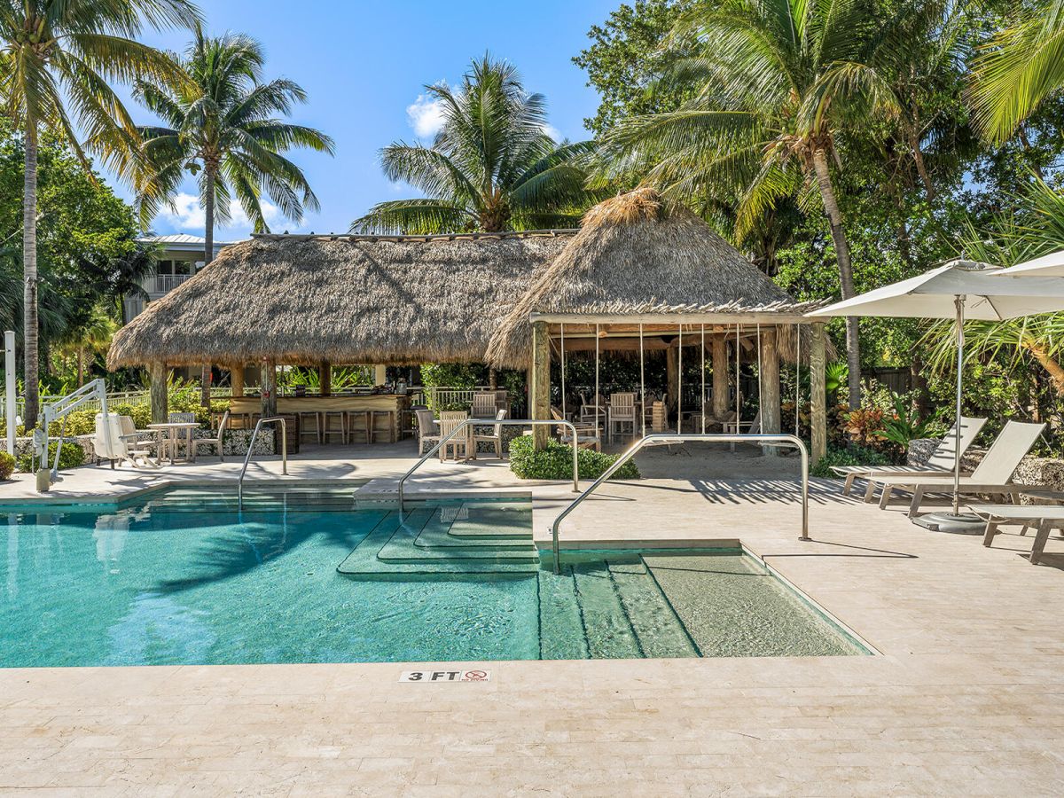 A tropical pool area with a thatched-roof bar, surrounded by palm trees and lounge chairs under umbrellas.