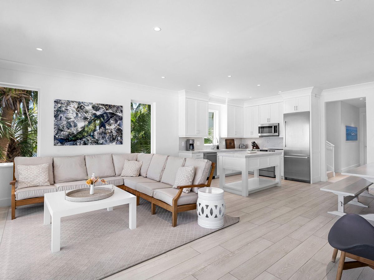 A modern living room with a neutral color scheme includes a sofa, coffee table, kitchen area, and wall art, creating an open and airy space.