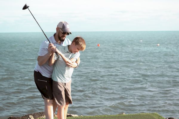 A person guides a child in swinging a golf club on a grass platform by the sea, under a clear sky.