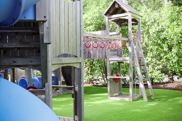 The image shows a playground with a blue slide, wooden climbing structures, and green artificial turf, surrounded by trees.