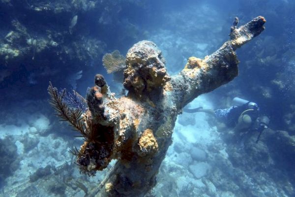 A coral-encrusted underwater statue with outstretched arms, accompanied by a scuba diver exploring the ocean surroundings.