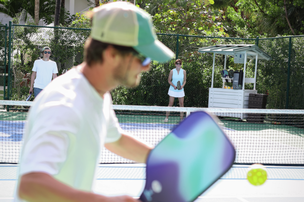 A person is playing pickleball on an outdoor court, holding a paddle, with a ball in motion, while others watch in the background.