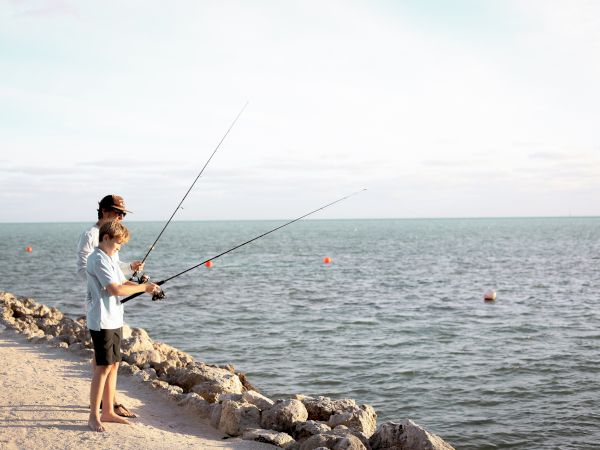 Two people are fishing by a rocky shoreline, facing a calm sea with fishing rods, under a clear sky with some clouds.