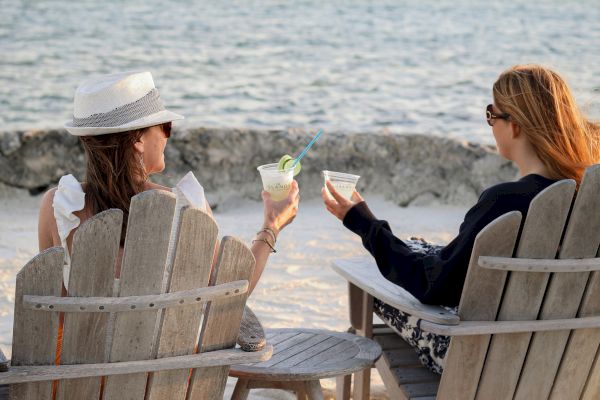 Two people sit in wooden chairs by the beach, holding drinks, enjoying the view and relaxation together.
