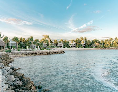 A scenic coastal view with palm trees, houses, and a calm sea under a blue sky with clouds, along a rocky shoreline.