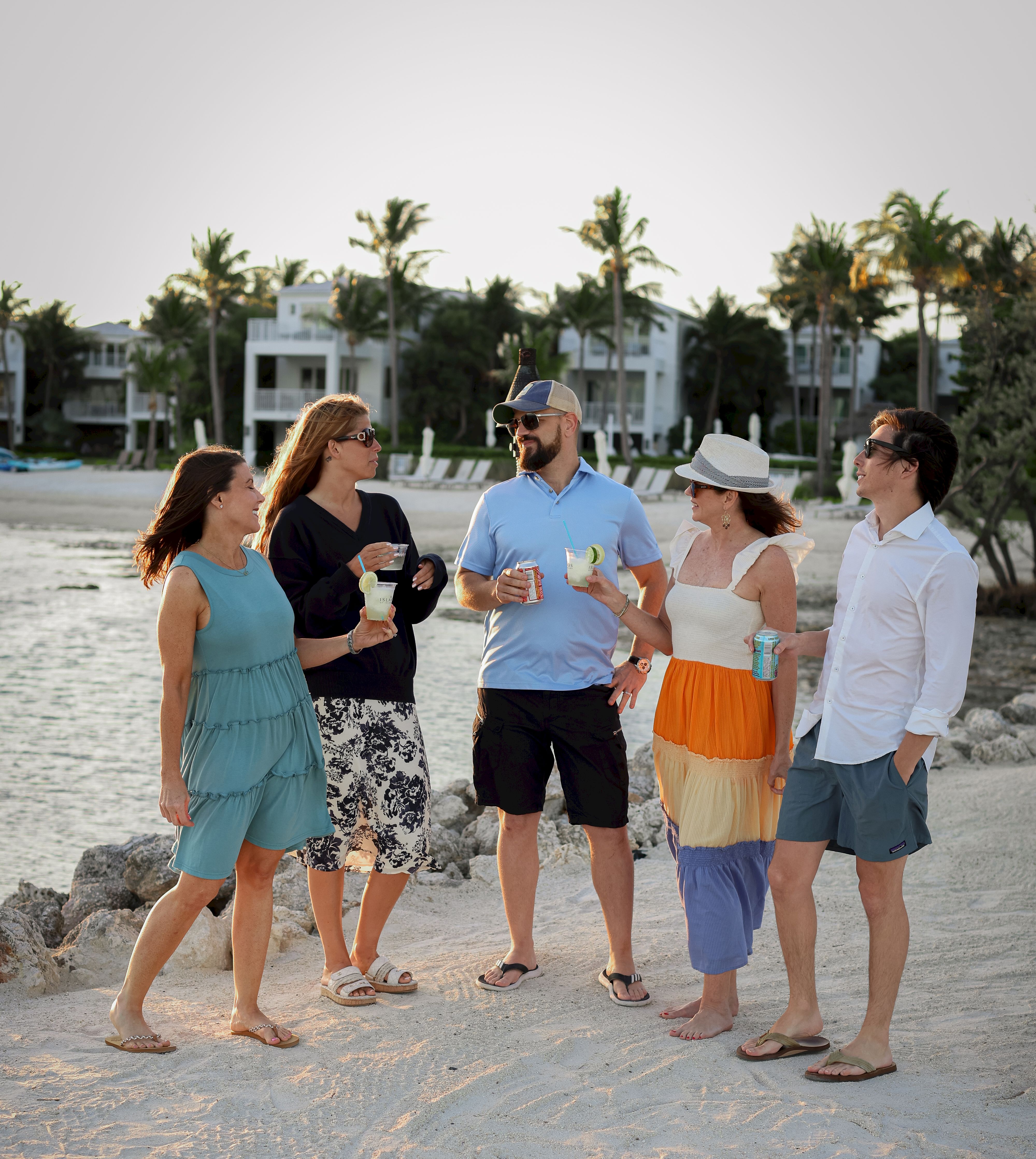 A group of people stand on a beach, chatting and holding drinks, with palm trees and buildings in the background.