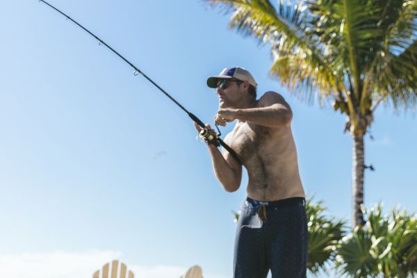 A person is fishing on rocky terrain with a palm tree and Adirondack chairs in the background under a clear blue sky.
