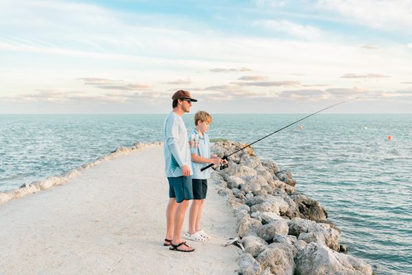 Two people are fishing on a rocky pier, surrounded by calm blue water under a partly cloudy sky.