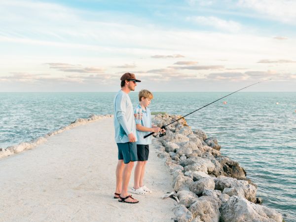 Two people are fishing on a rocky pier, surrounded by calm blue water under a partly cloudy sky.