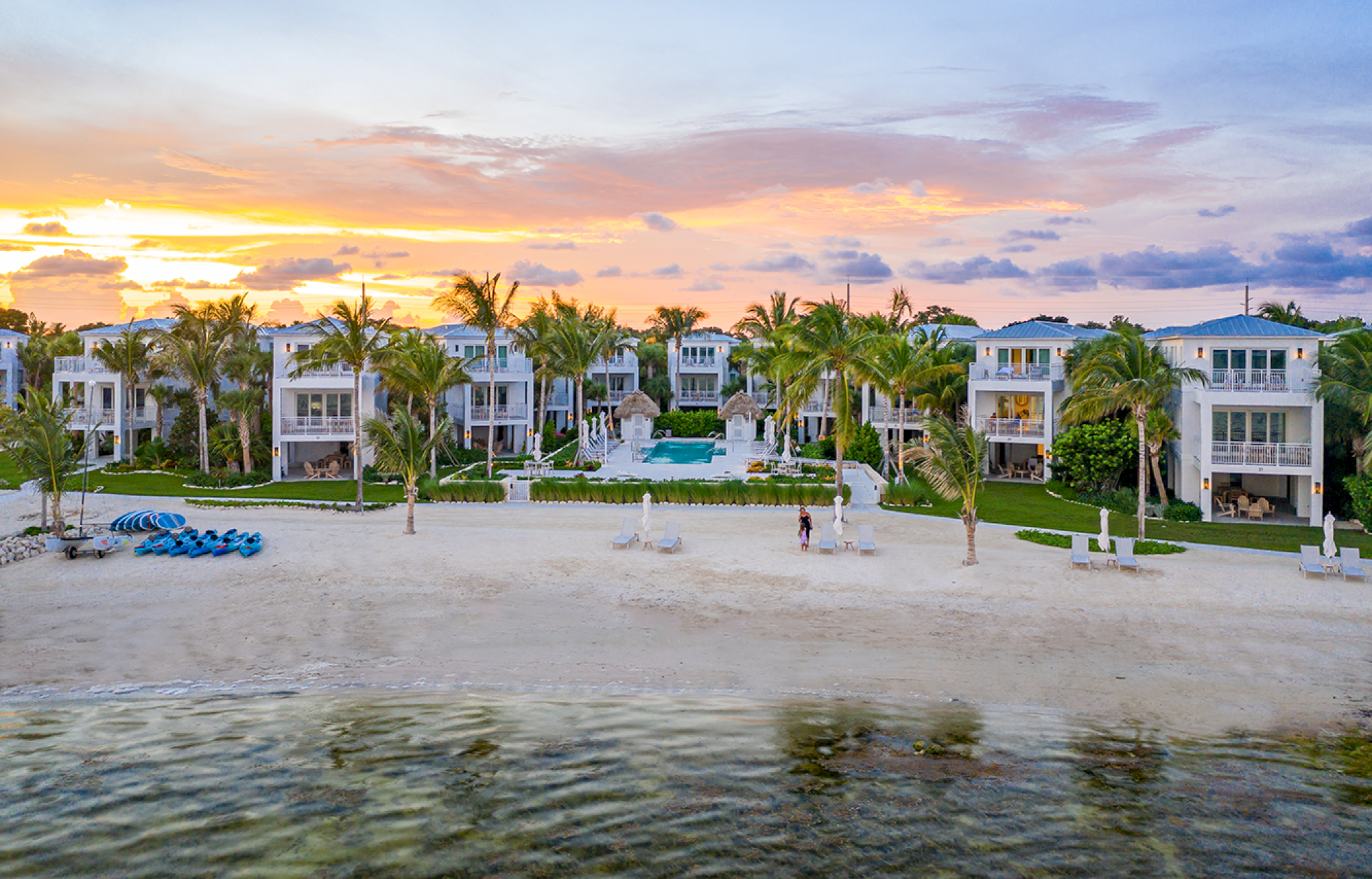 Beachfront view of luxury villas with palm trees, a sandy beach, and a sunset sky in the background.