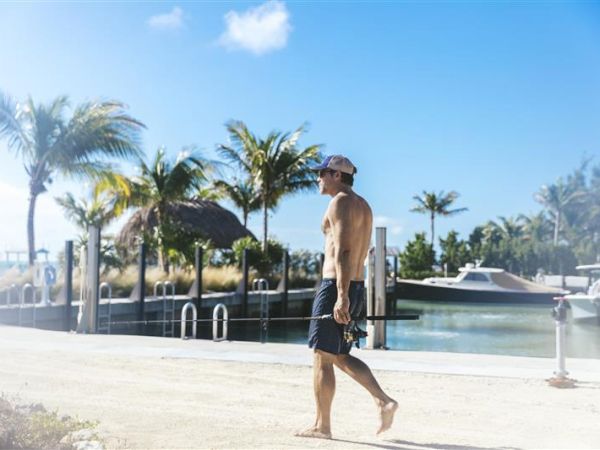 A person walks along a sunny waterfront with palm trees, a dock, and boats in the background, under a clear blue sky.