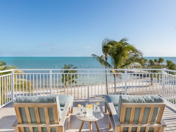 A balcony with two chairs and a table overlooks a sandy beach and calm ocean, framed by palm trees under a clear blue sky.