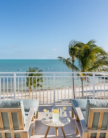 A beachside patio with two wooden chairs and a small table overlooks the ocean, surrounded by palm trees under a clear blue sky.