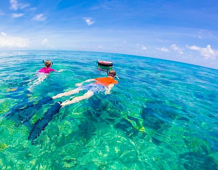 Two people snorkel in clear turquoise ocean near a small boat on a sunny day, exploring vibrant coral beneath the surface.